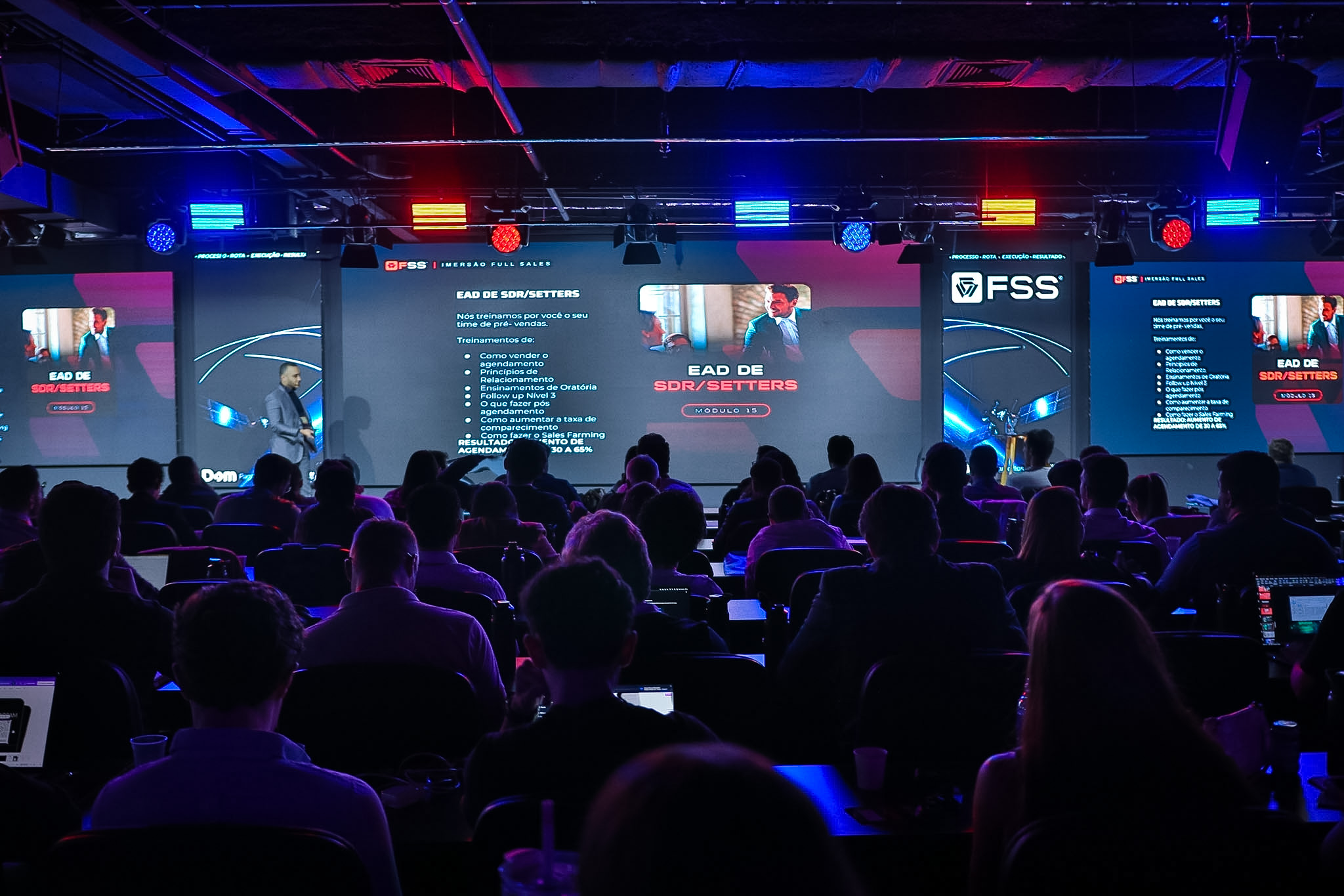 Palco com 3 telões em uso durante palestra com iluminação cênica