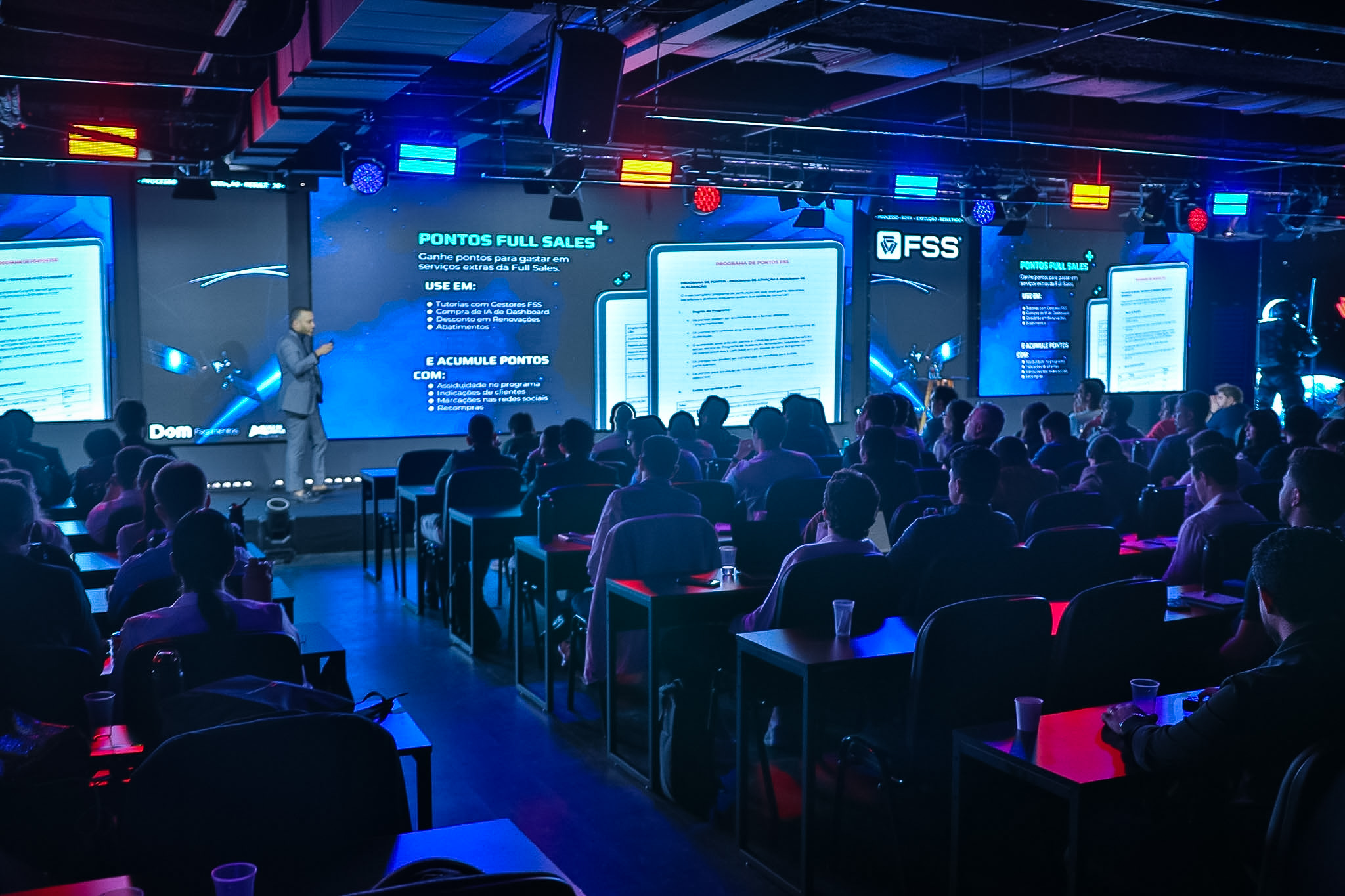 Palestrante no palco com microfone e telões exibindo conteúdo